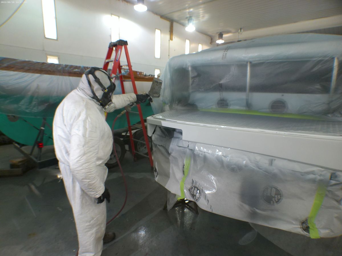 A person in protective gear is spray painting or coating a boat in a workshop. The boat and nearby areas are covered with protective plastic sheeting.