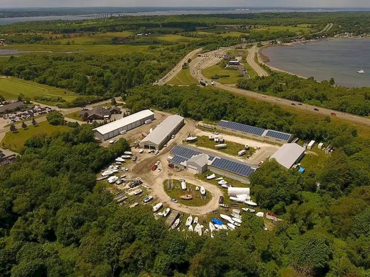 An aerial view of a rural facility with multiple buildings and parked boats, surrounded by greenery, near a coastline with a road running along it.