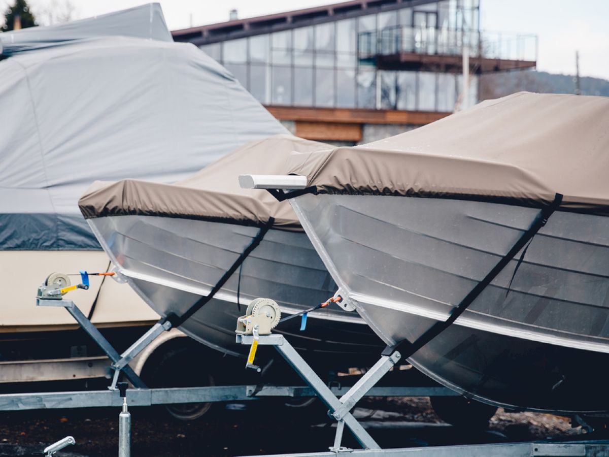 The image shows two covered boats on trailers, positioned side by side in an outdoor storage area with a modern building in the background.