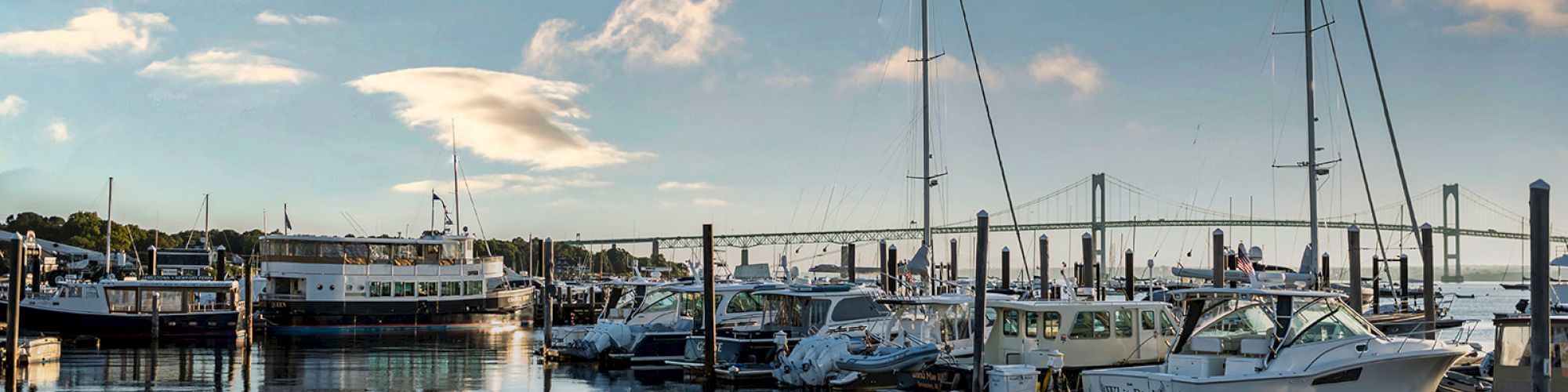 The image shows a marina with boats docked, calm water reflecting clouds, and a bridge in the background under a partly cloudy sky.