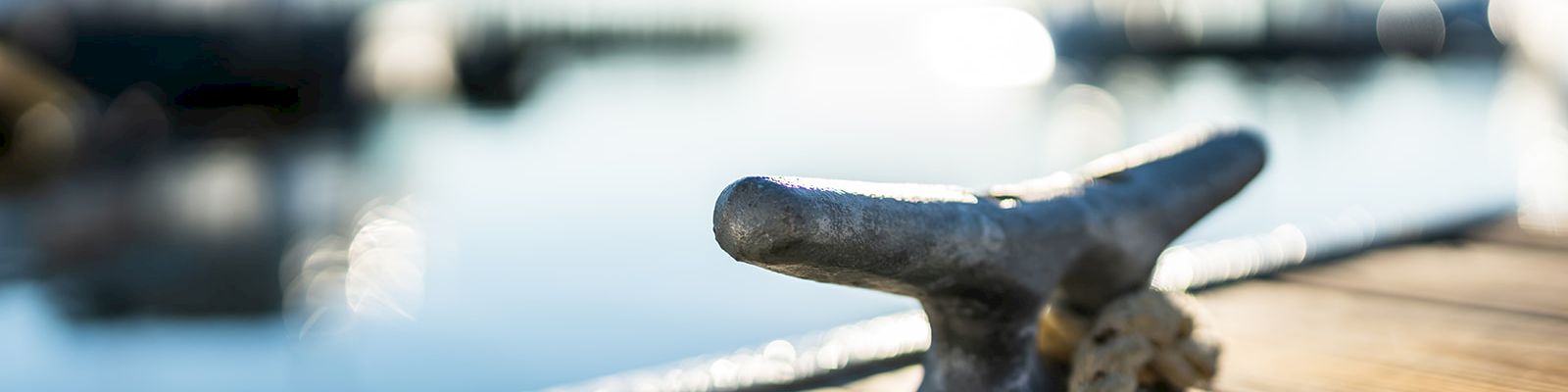 A close-up of a dock cleat with a mooring rope on a wooden pier, with a blurred boat and harbor in the background.