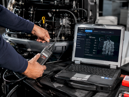 A mechanic plugs a Yamaha diagnostic tool into an engine while a laptop displays live test data.