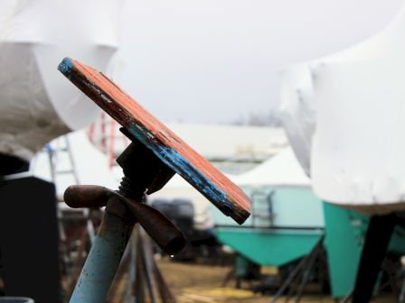 A close-up of a chipped wooden plank with a rusty metal pipe in a workshop yard, with tools and materials blurred behind.