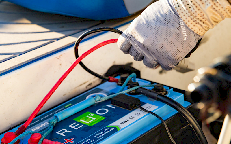 A gloved technician connects red/black cables to a blue battery pack on a vehicle or boat, with wires and tools nearby.