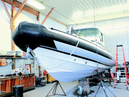 A boat sits on a workshop stand under a hangar roof, with a long black hull section and tools/ladders nearby.