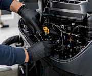 A mechanic in gloves works inside a car’s engine bay, adjusting hoses and parts under the hood.