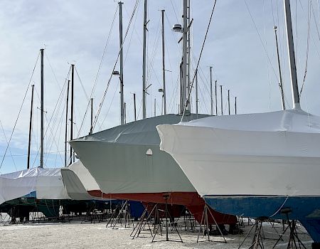 A row of sailboats on launch stands, covered with white protective tarps, at a dock/marina under a cloudy sky.