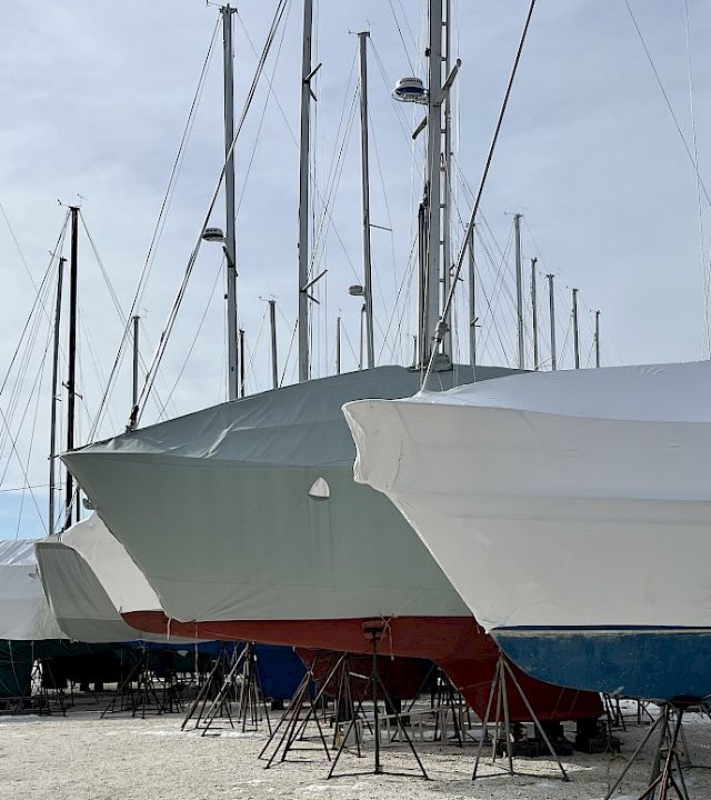 A row of sailboats on launch stands, covered with white protective tarps, at a dock/marina under a cloudy sky.