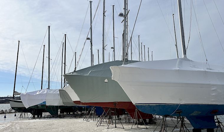 A row of sailboats on launch stands, covered with white protective tarps, at a dock/marina under a cloudy sky.