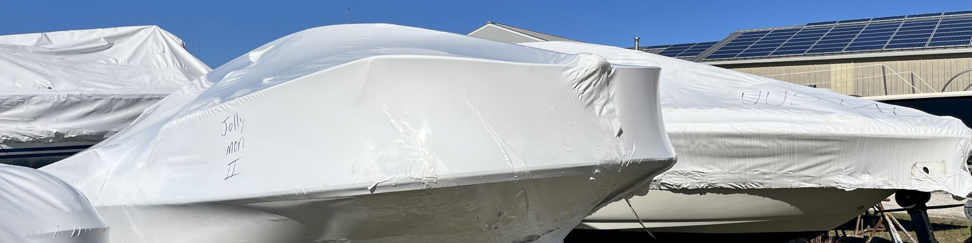 A white jet is covered in protective white tarp on a trailer outdoors, with solar panels and a blue sky in the background.