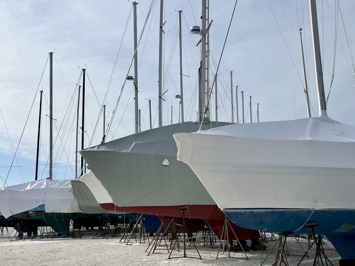 Sailboats on maintenance stands with tall masts and protective white covers, moored by the water under a cloudy sky.