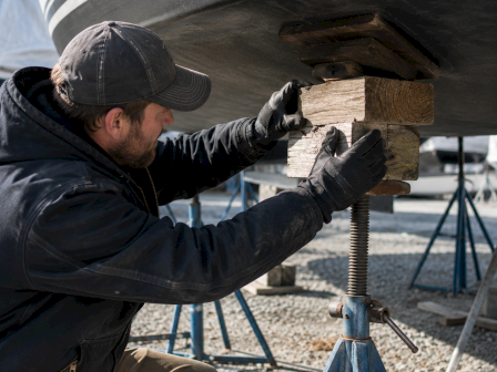 A worker in gloves repairs wood on a boat hull with tools, wearing a cap and crouching on a stand.