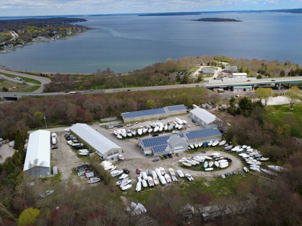 Aerial view of a riverside marina with many boats, solar‑panel buildings, and a nearby bridge over the water.