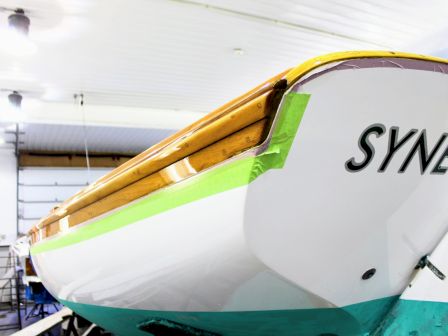 A yellow-and-white plane fuselage inside a hangar, with green tape over the cockpit area and &ldquo;SYNEC&rdquo; on the side.