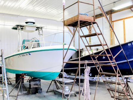 An indoor workshop with boats on stands, scaffolding and a ladder, showing a white-and-green boat/jet ski and a blue boat.