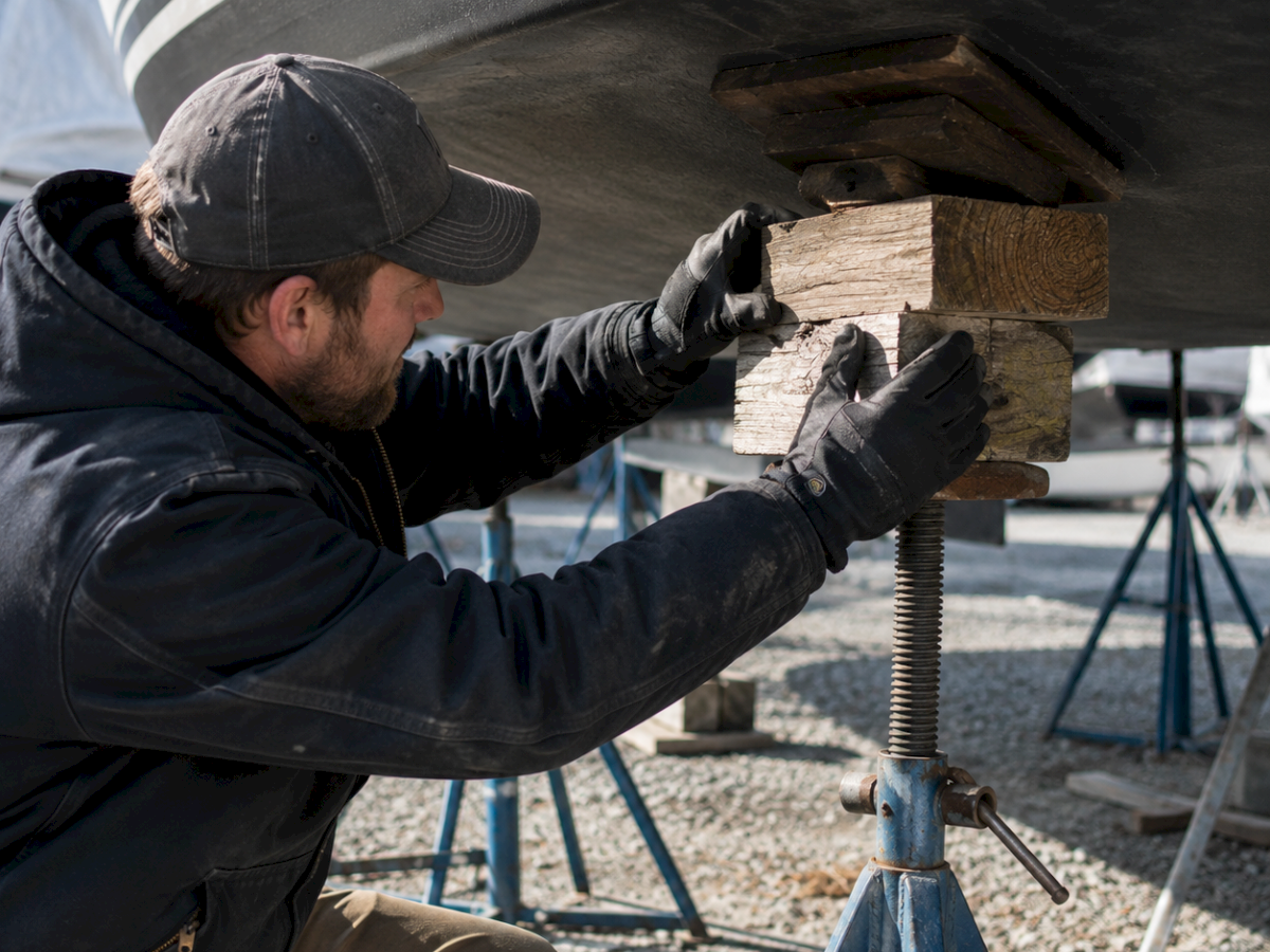 A worker in gloves and a cap installs or repairs wooden planks, kneeling under a large ship-like structure.