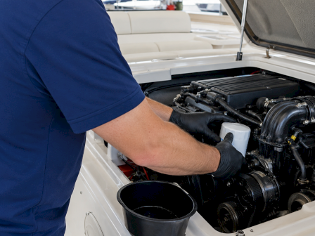 A mechanic in blue coveralls wears gloves while working inside a car&rsquo;s open engine bay near the hood.