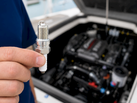 A person holds a silver spark plug while working on a car engine bay filled with tools.