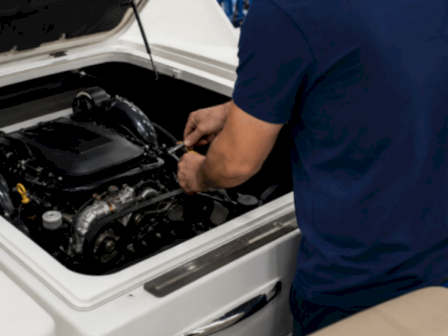 A mechanic in blue works on a car engine under the open hood, tightening a part with a tool.