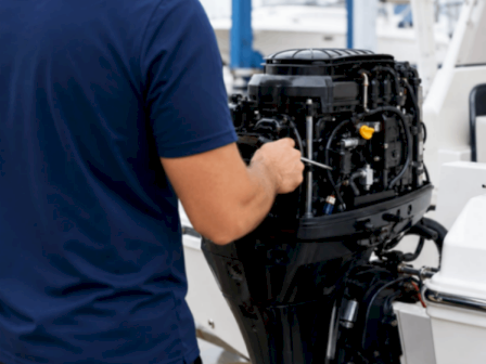 A technician in a blue shirt works on a boat&rsquo;s outboard engine in a workshop.
