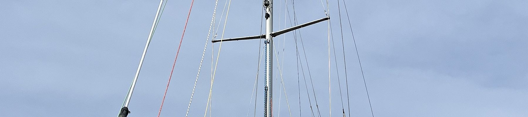 A white sailboat with tall mast and rigging mounted on supports, with the sky overhead and trees by the waterfront.