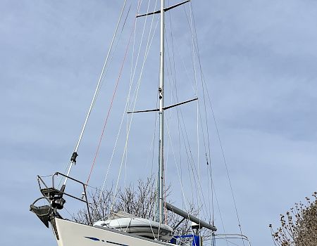 A white sailboat with tall mast and rigging mounted on supports, with the sky overhead and trees by the waterfront.