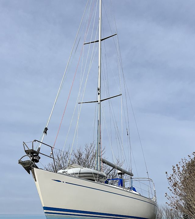 A white sailboat with tall mast and rigging mounted on supports, with the sky overhead and trees by the waterfront.