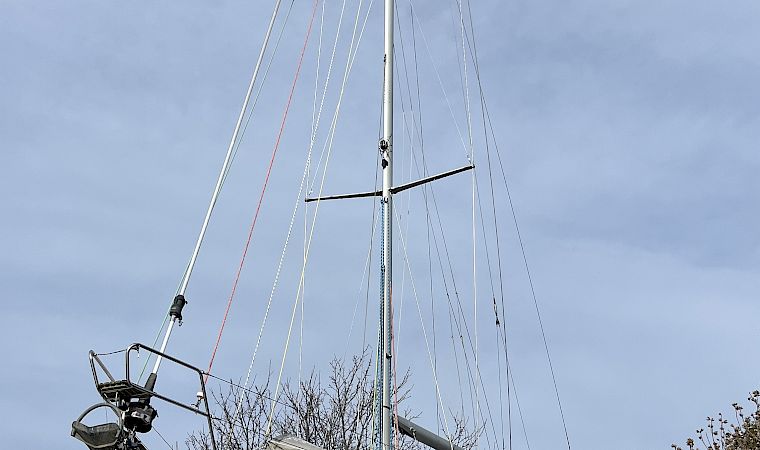 A white sailboat with tall mast and rigging mounted on supports, with the sky overhead and trees by the waterfront.
