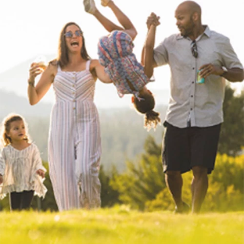 A family enjoys a sunny day outdoors, with the parents lifting a child high while another child watches and smiles. Everyone appears to be happy.
