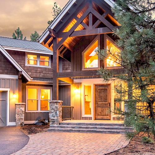 This image shows a large, well-lit house with a rustic design, featuring two garage doors, wooden elements, and surrounded by pine trees.