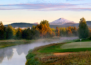 A scenic view of a golf course with a river, trees, and a snow-capped mountain in the background during sunrise or sunset.