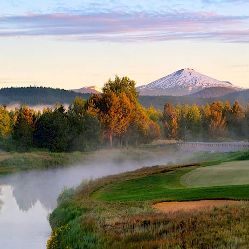 A scenic view of a golf course with a river, trees, and a snow-capped mountain in the background during sunrise or sunset.