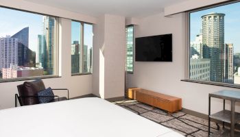A modern room with a large bed, armchair, flat-screen TV, two large windows showcasing a cityscape with high-rise buildings, ending the sentence.