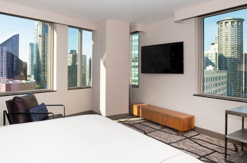 A modern room with a large bed, armchair, flat-screen TV, two large windows showcasing a cityscape with high-rise buildings, ending the sentence.