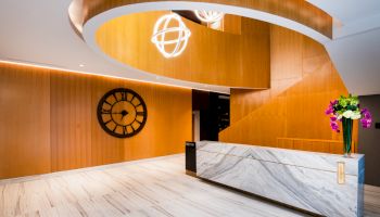 A modern lobby area with a curved wooden staircase, marble reception desk, large wall clock, and decorative lighting fixtures, and flowers on the desk.