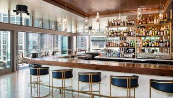 A modern bar with a wooden counter, high chairs, and a wide assortment of bottles on shelves in a well-lit, open space with city views.