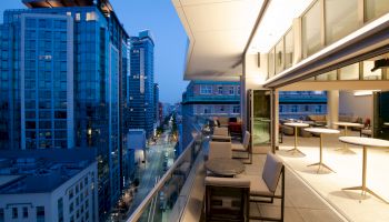 The image shows a modern cityscape at dusk with tall buildings and a furnished balcony that includes tables and chairs on the right.