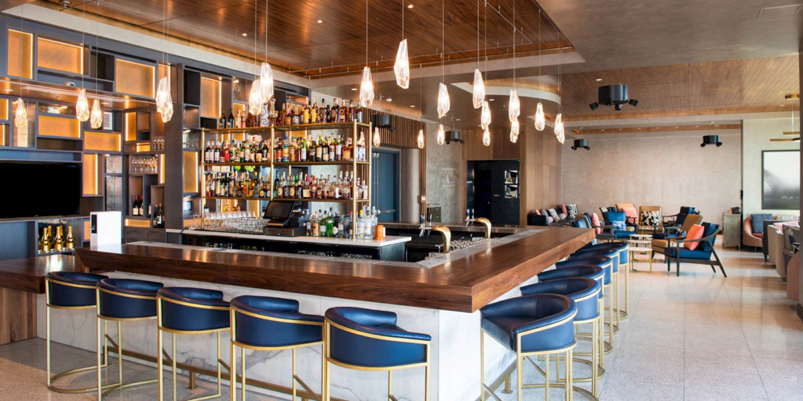 The image shows a modern bar interior with a wooden counter, blue stools, hanging lights, a well-stocked liquor shelf, and a seating area.