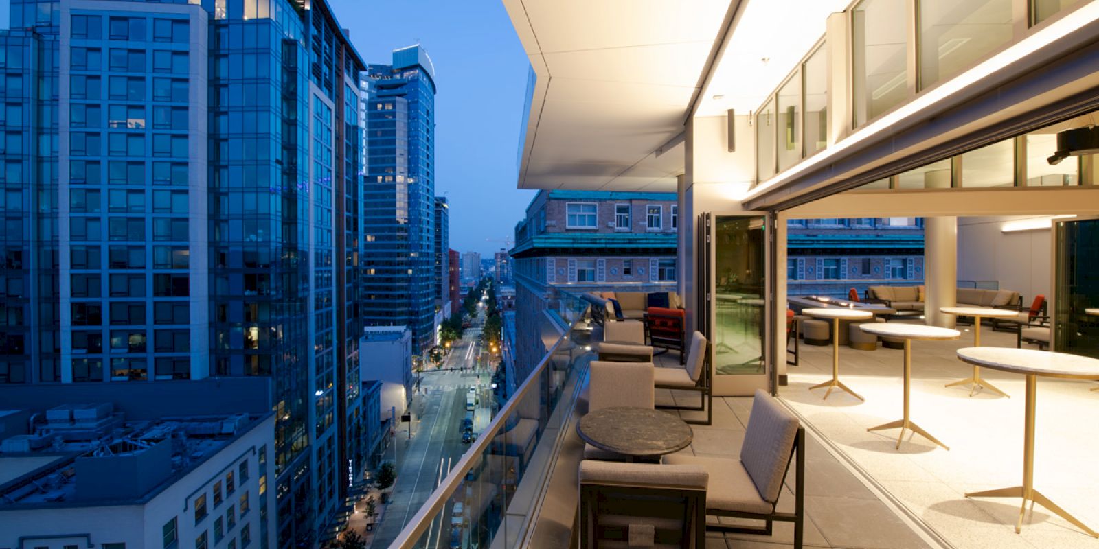 A modern balcony with seating and tables overlooks a cityscape at dusk, lined with high-rise buildings and lit streets.