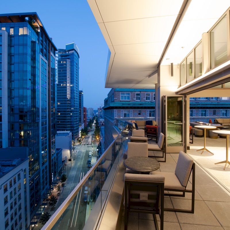 A modern cityscape at dusk with a view from a high-rise balcony featuring outdoor seating and tables on the terrace.