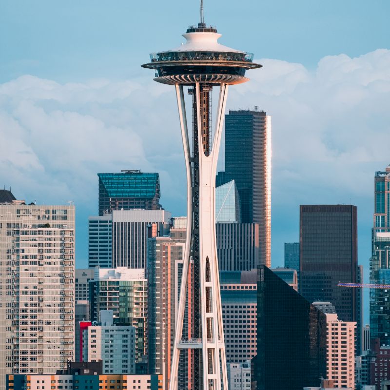 The image shows the Space Needle tower located in Seattle, Washington, with a backdrop of various skyscrapers and buildings under a cloudy sky.