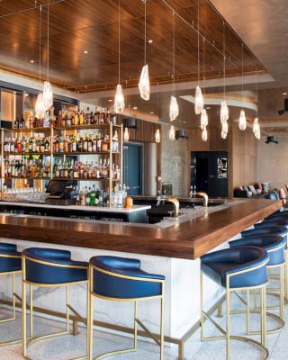 The image shows a modern bar with a wooden counter, blue bar stools, hanging lights, and a well-stocked back wall. There is seating in the background.