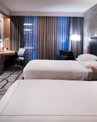 A modern hotel room with two neatly made beds, a wall-mounted TV, a desk and chair, ambient lighting, and a floor-to-ceiling window with curtains.