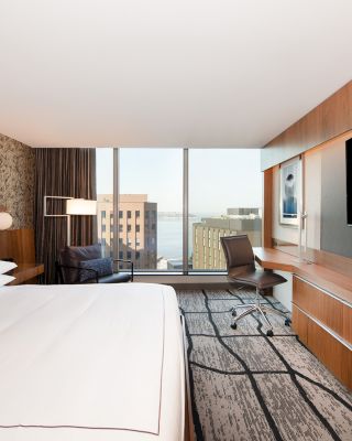 The image shows a modern hotel room with a large bed, a desk and chair, a flat-screen TV, and a window with a cityscape view.