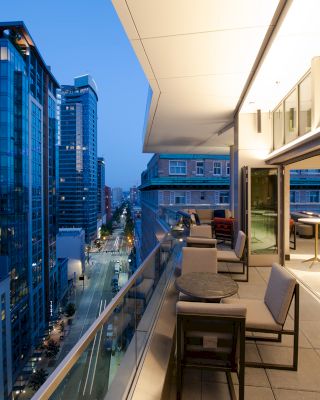 This image shows a modern balcony at twilight overlooking a city street lined with tall buildings, with seating and tables present on the balcony.