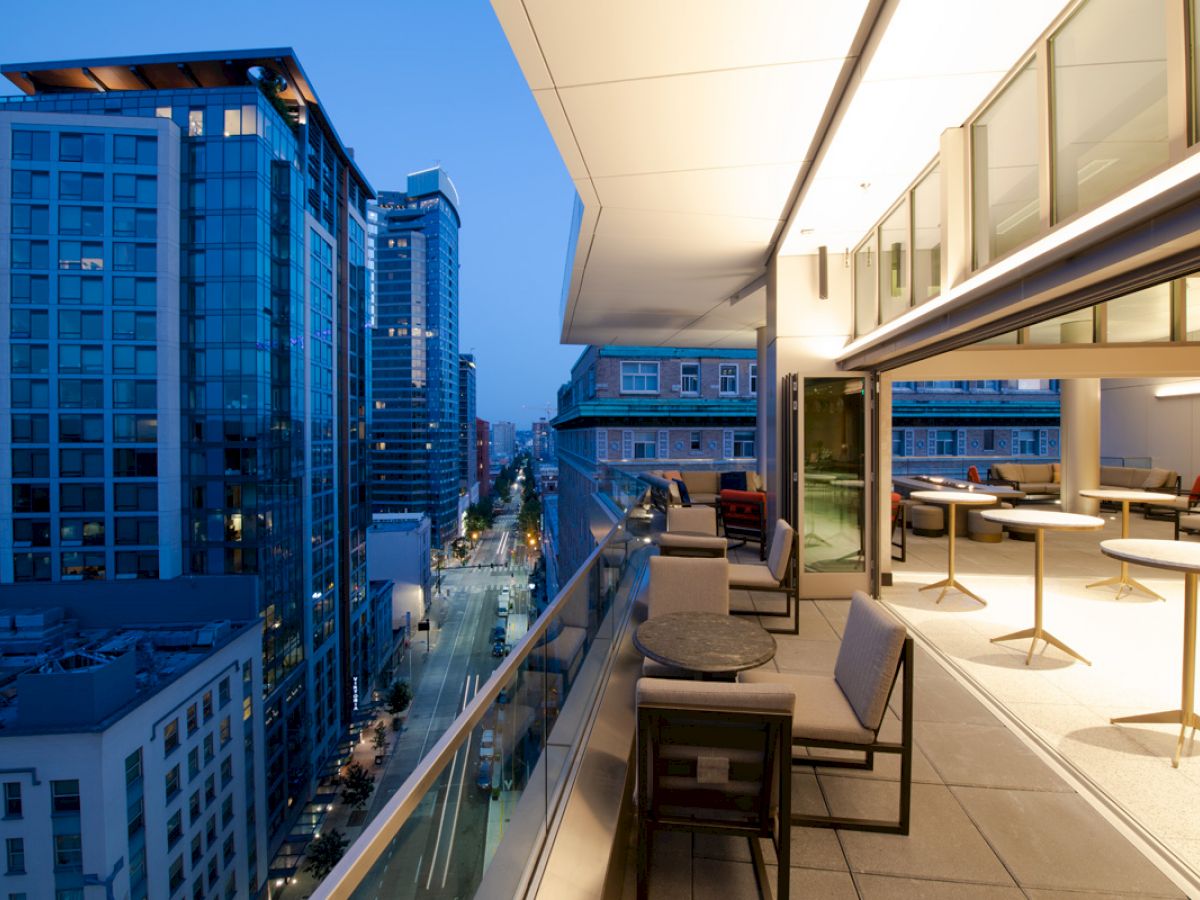 A modern high-rise balcony at dusk, with outdoor seating and tables, overlooks a city street lined with tall buildings.