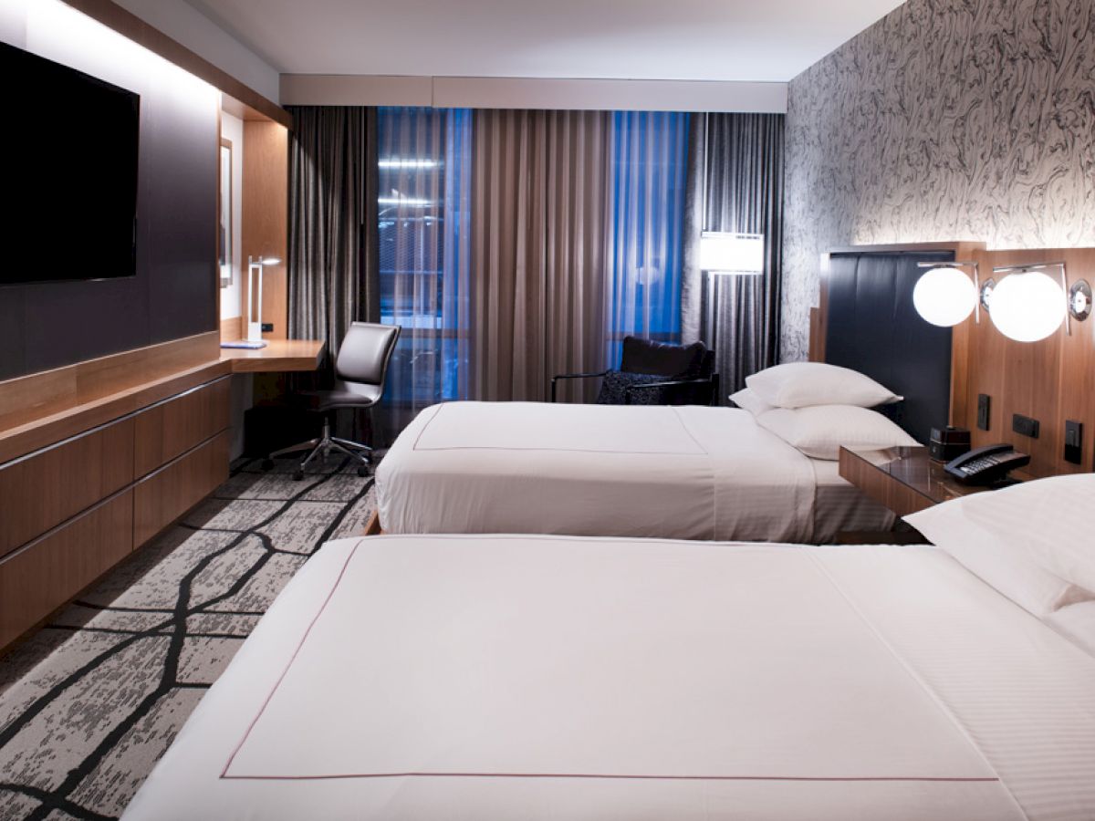 A modern hotel room with two double beds, a wall-mounted TV, a work desk by the window, and contemporary decor.