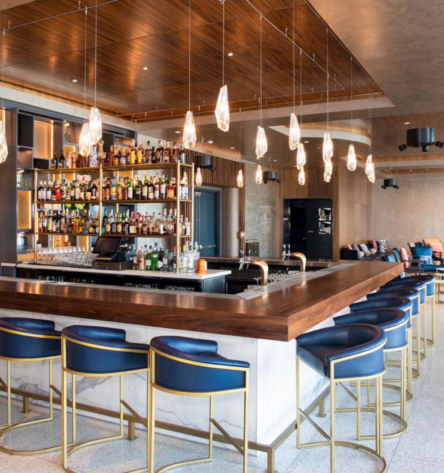 A modern bar with a wooden counter, blue stools, an array of bottles, hanging lights, and lounge seating in the background ending the sentence.