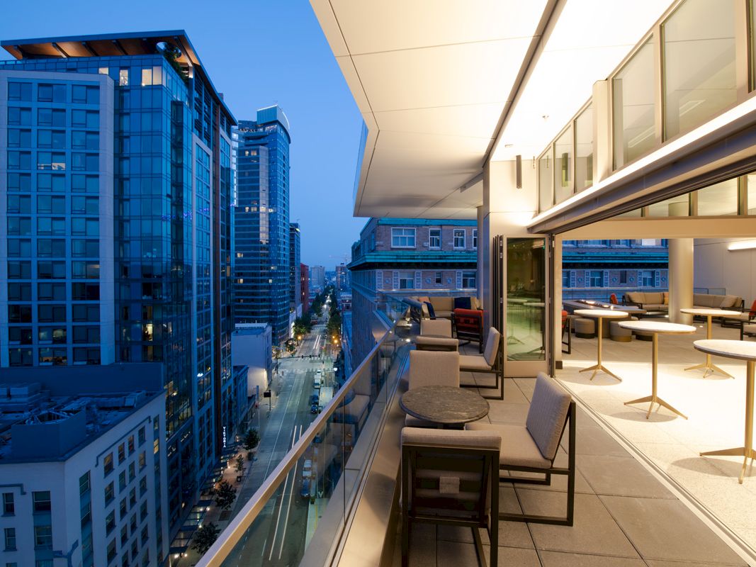 A modern cityscape at dusk with high-rise buildings, a well-lit balcony featuring tables and chairs overlooking a busy street.