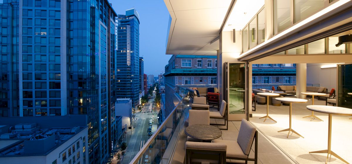 A modern cityscape at dusk with high-rise buildings, a well-lit balcony featuring tables and chairs overlooking a busy street.